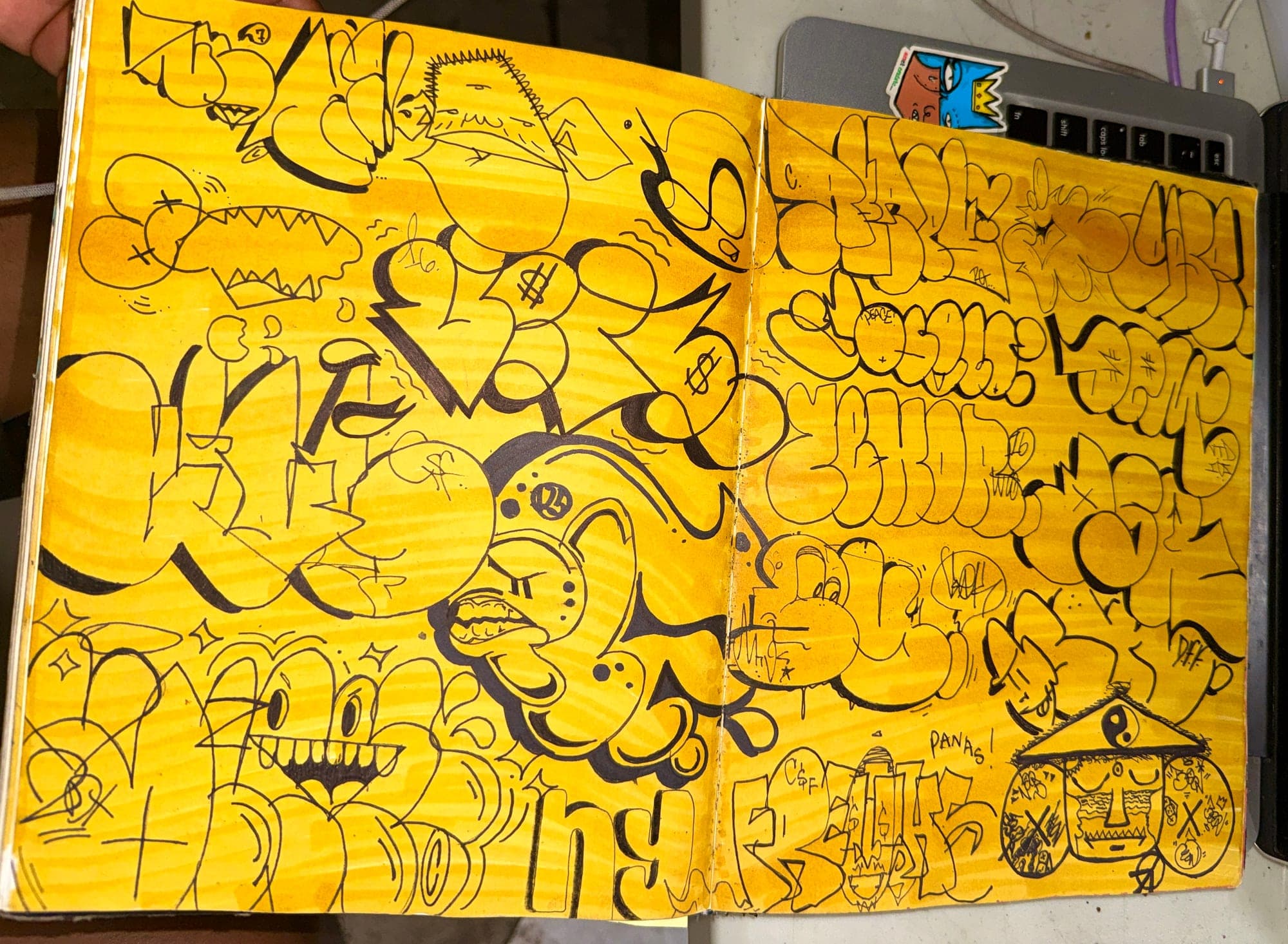 A blackbook open to a two-page spread of black marker throw-ups and characters over yellow
