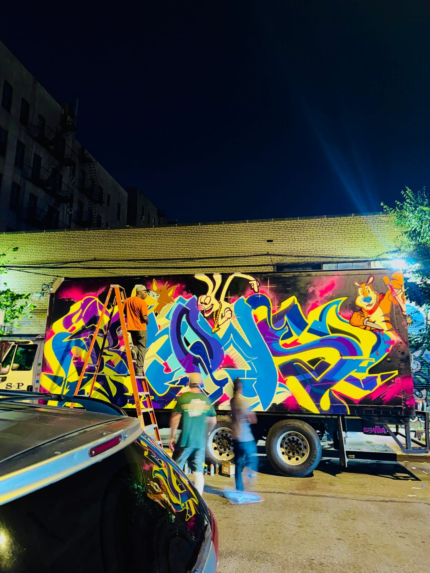 A box truck being painted at night, one writer up an orange ladder while people walk past