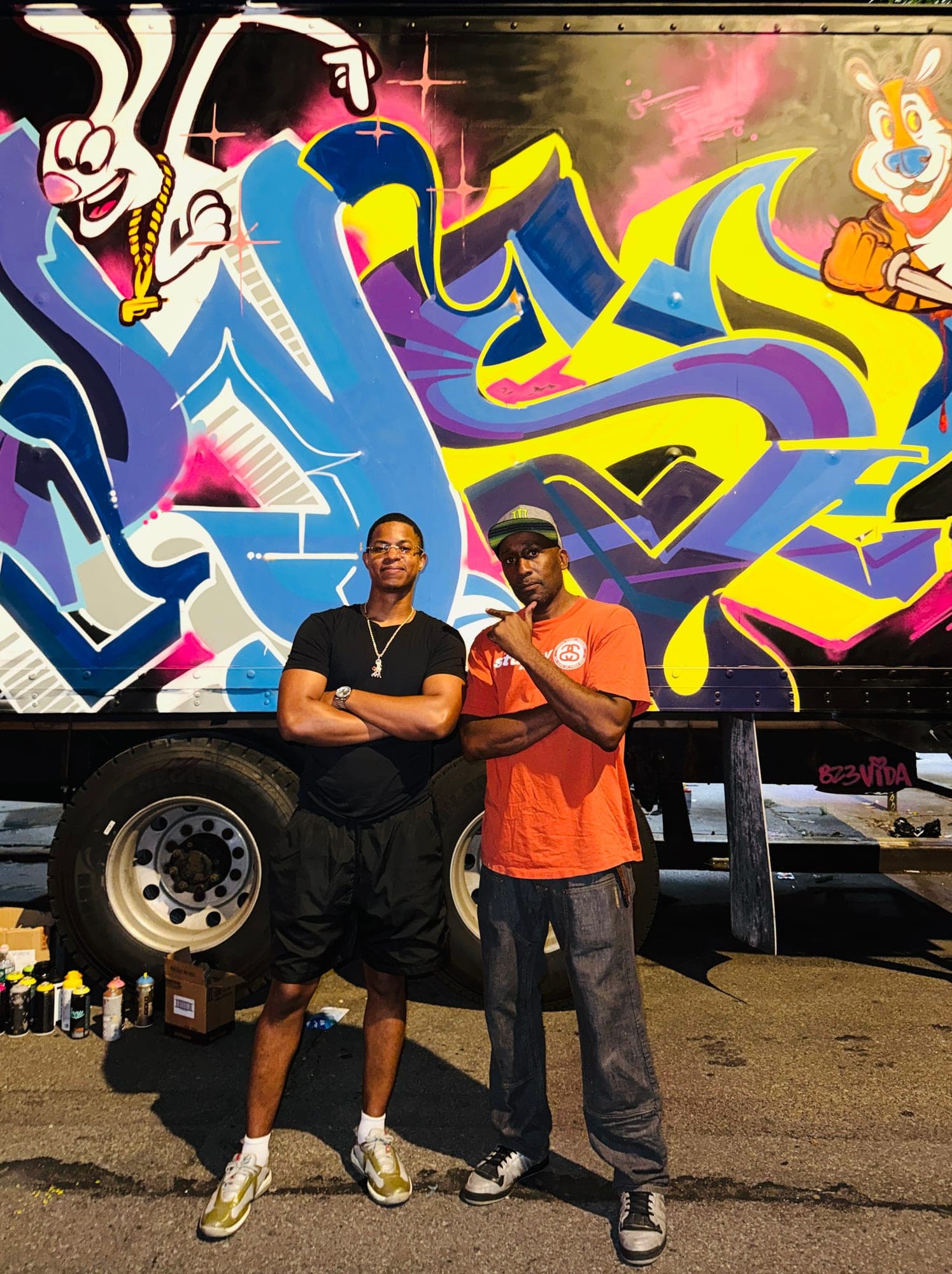 Two men standing in front of the painted box truck, spray cans lined up on the ground beside them