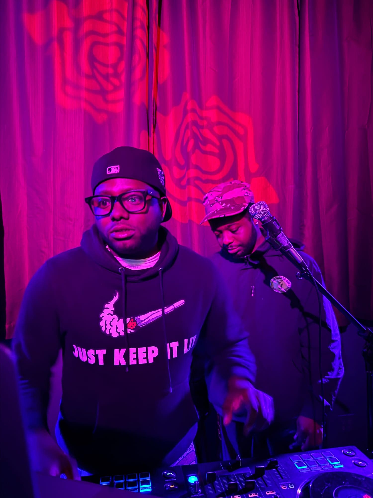 A DJ in glasses and a cap behind a controller, a second man at the mic, roses projected on the red curtain behind them