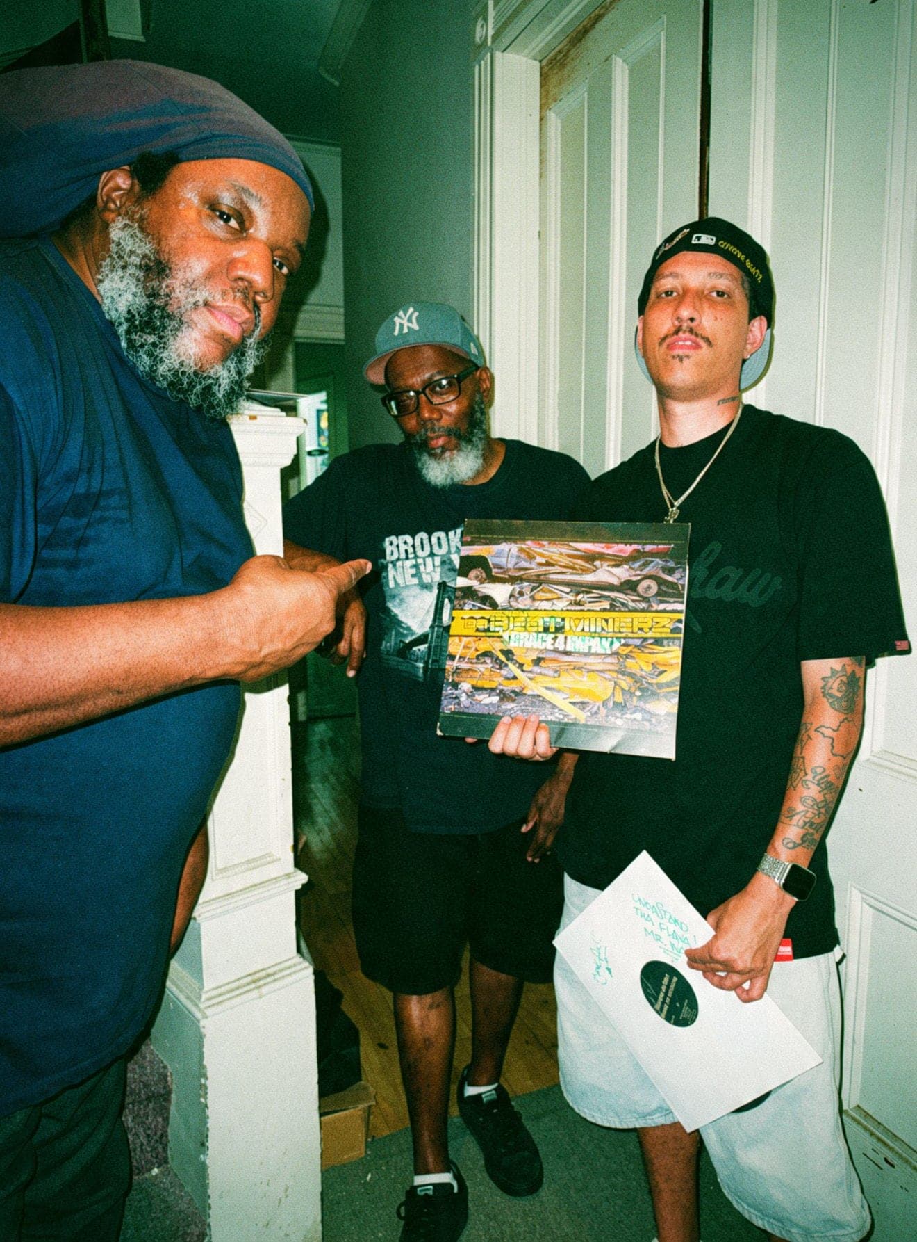 Three men in a hallway, one holding up a record sleeve and another a white-label twelve-inch