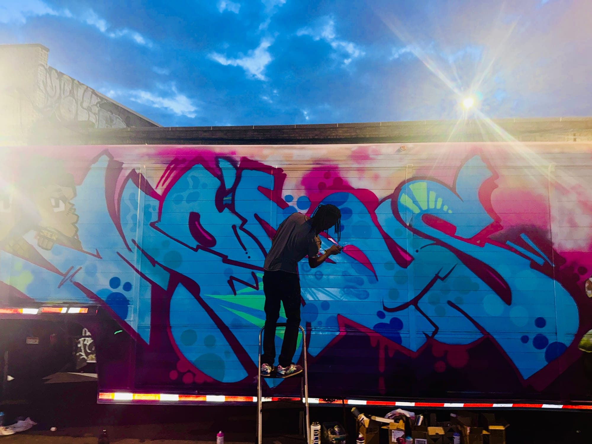A writer up a stepladder painting a big blue and magenta piece along the side of a trailer at dusk