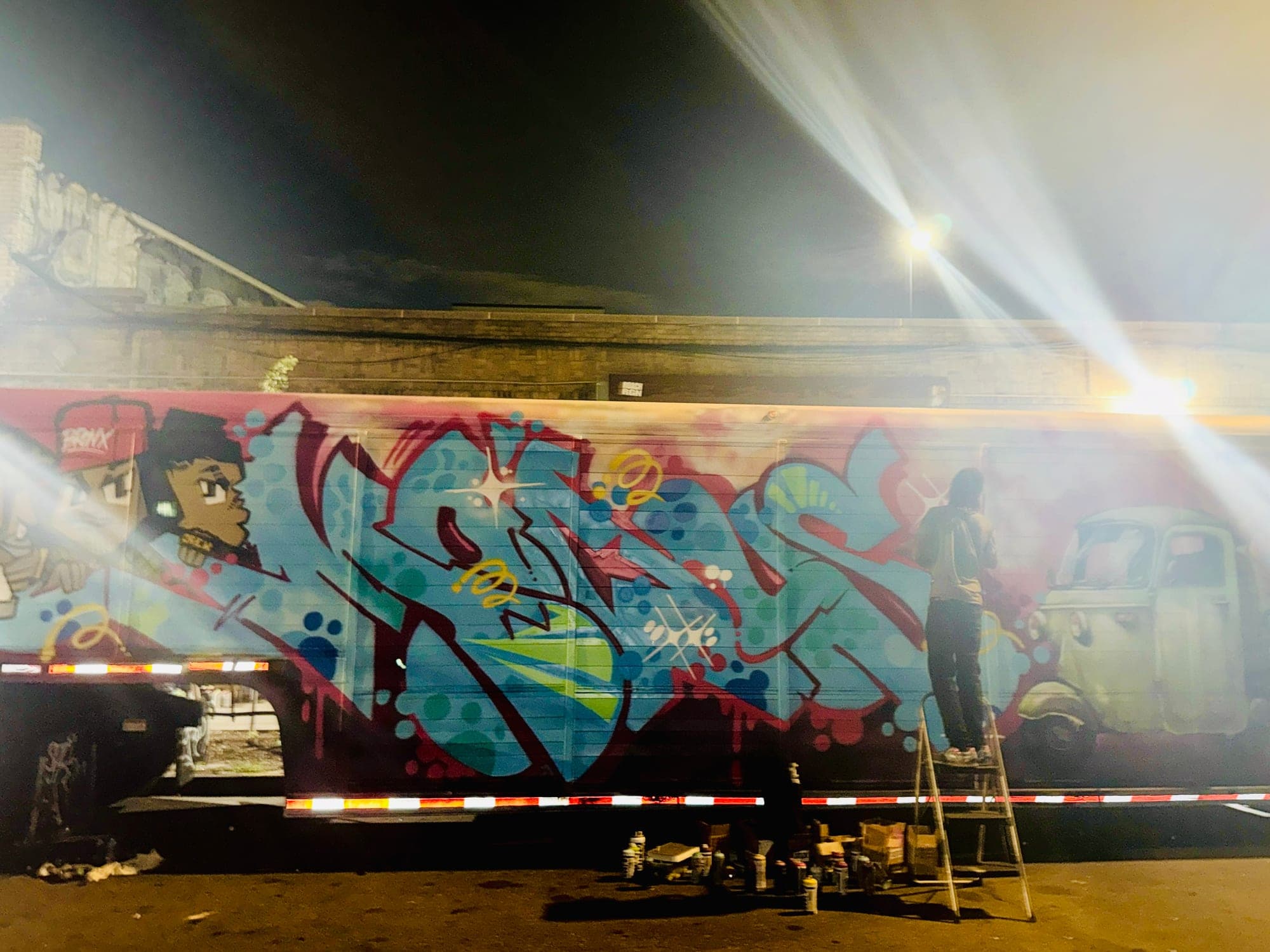 The same trailer piece after dark under work lights, a character painted at one end and a pickup truck at the other